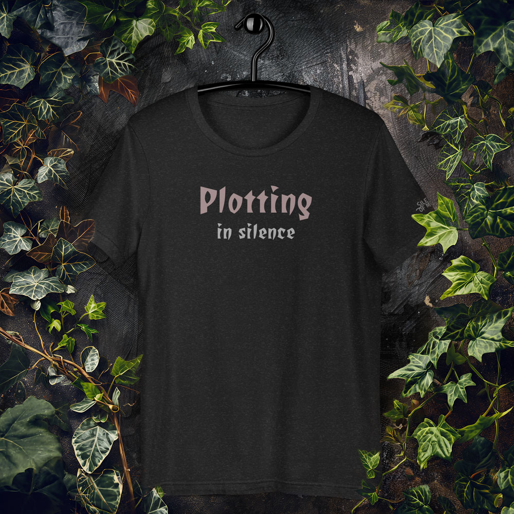 Gothic-style text design reading “Plotting in Silence” in bold, moody font on a black t-shirt.